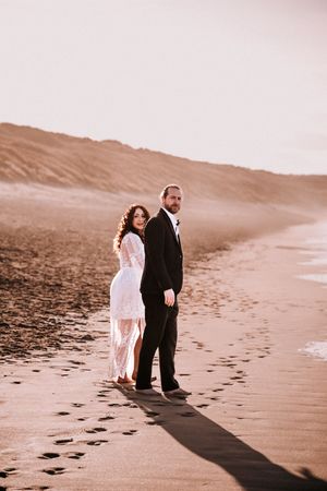 A couple in wedding attire walks along a serene beach at sunset leaving footprints in the sand.
