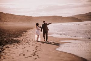 Two people walking along a sepia-toned beach shoreline leaving footprints in the sand at sunset.