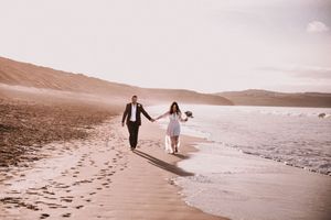 A couple in wedding attire walks hand in hand along a serene beach at sunset with mountains in the background.