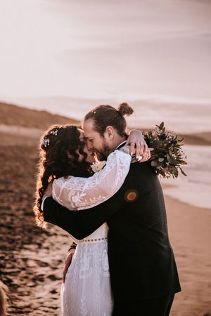 A couple in wedding attire embraces passionately on a beach during golden hour.
