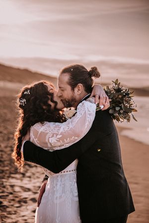 A couple in wedding attire embracing on a beach at sunset with golden light behind them.