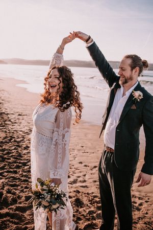 A couple dances joyfully together on a sandy beach at sunset in romantic wedding attire.