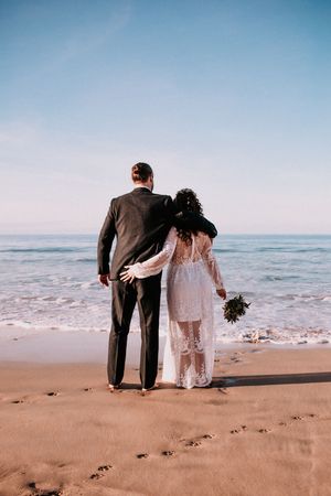 A bride in lace and groom in suit walk together on a sandy beach with ocean waves in background.