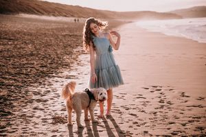 A person in a blue dress plays with a fluffy dog on a sandy beach at sunset.