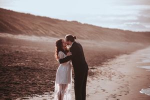 A tender moment between a couple in wedding attire on a moody beach at sunset.