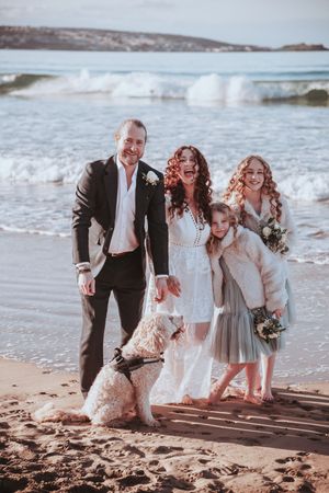 A wedding party poses on the beach with their dog as waves crash in the background.