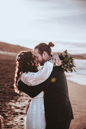 A romantic embrace between newlyweds on a beach at sunset with a bouquet of flowers.
