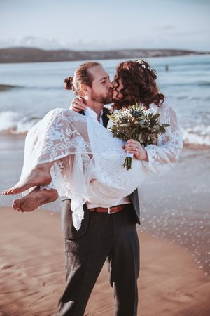 A romantic beach wedding photo of a couple embracing, with the bride holding a bouquet against an ocean backdrop.