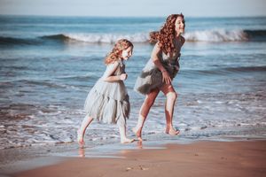 Two children in light dresses run joyfully along the shoreline as waves crash behind them. emotive wedding photography.
