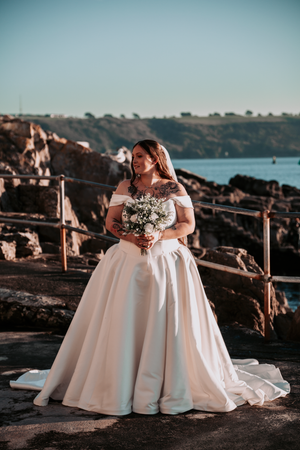 Wedding dress silhouette against coastal backdrop with white bouquet.