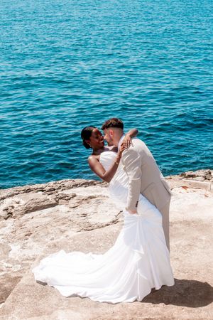 A couple in wedding attire shares a romantic moment on rocky cliffs overlooking bright blue waters.