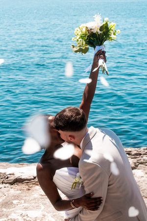 A couple embraces on coastal rocks with flower petals falling around them.