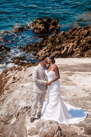 A romantic wedding couple poses on rocky coastal cliffs overlooking bright blue ocean waters.