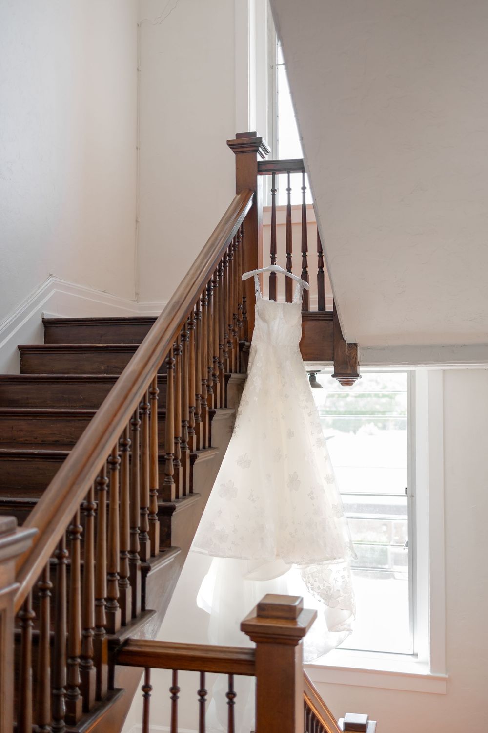 A flowing white wedding dress hangs elegantly against a wooden staircase with natural light coming through nearby windows. NASHVILLE WEDDING