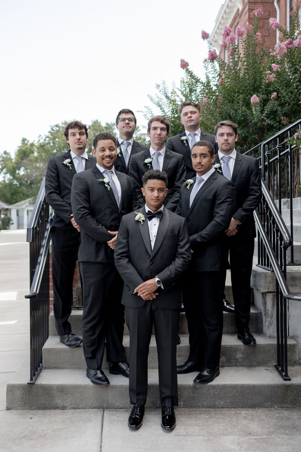 Groomsmen in black suits pose casually on outdoor stone steps with flowering bushes in the background. NASHVILLE WEDDING PHOTOGRAPHER