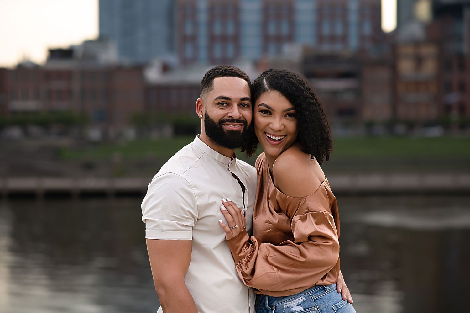 A casual engagement portrait session by a waterfront with urban architecture in the background.