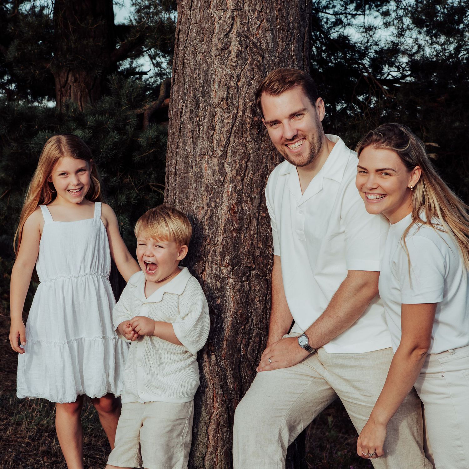 Natural photo of a Mum and dad with their 2 young children laughing by a tree in the evening sun.