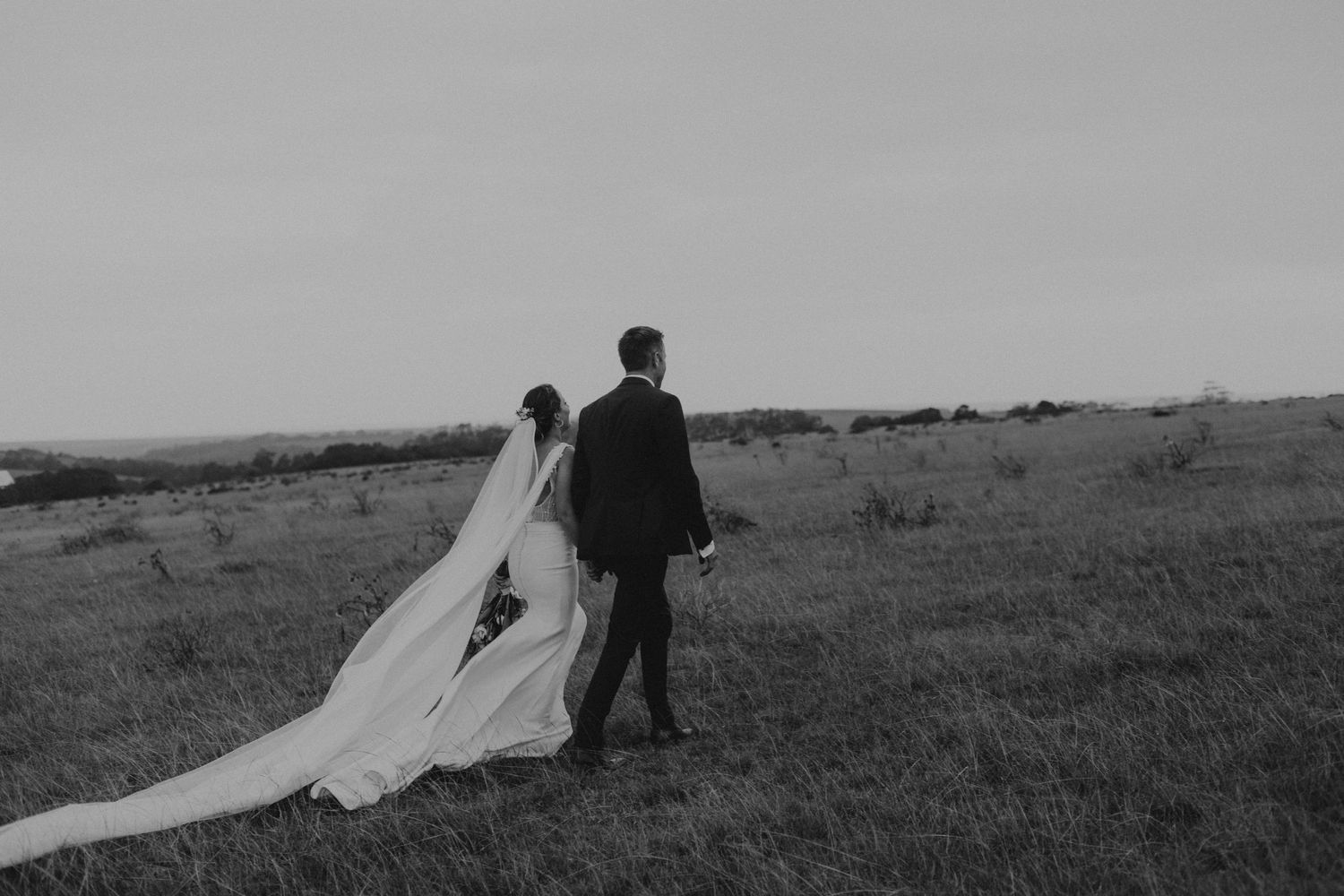 Couple walking through windswept field with flowing wedding dress veil.