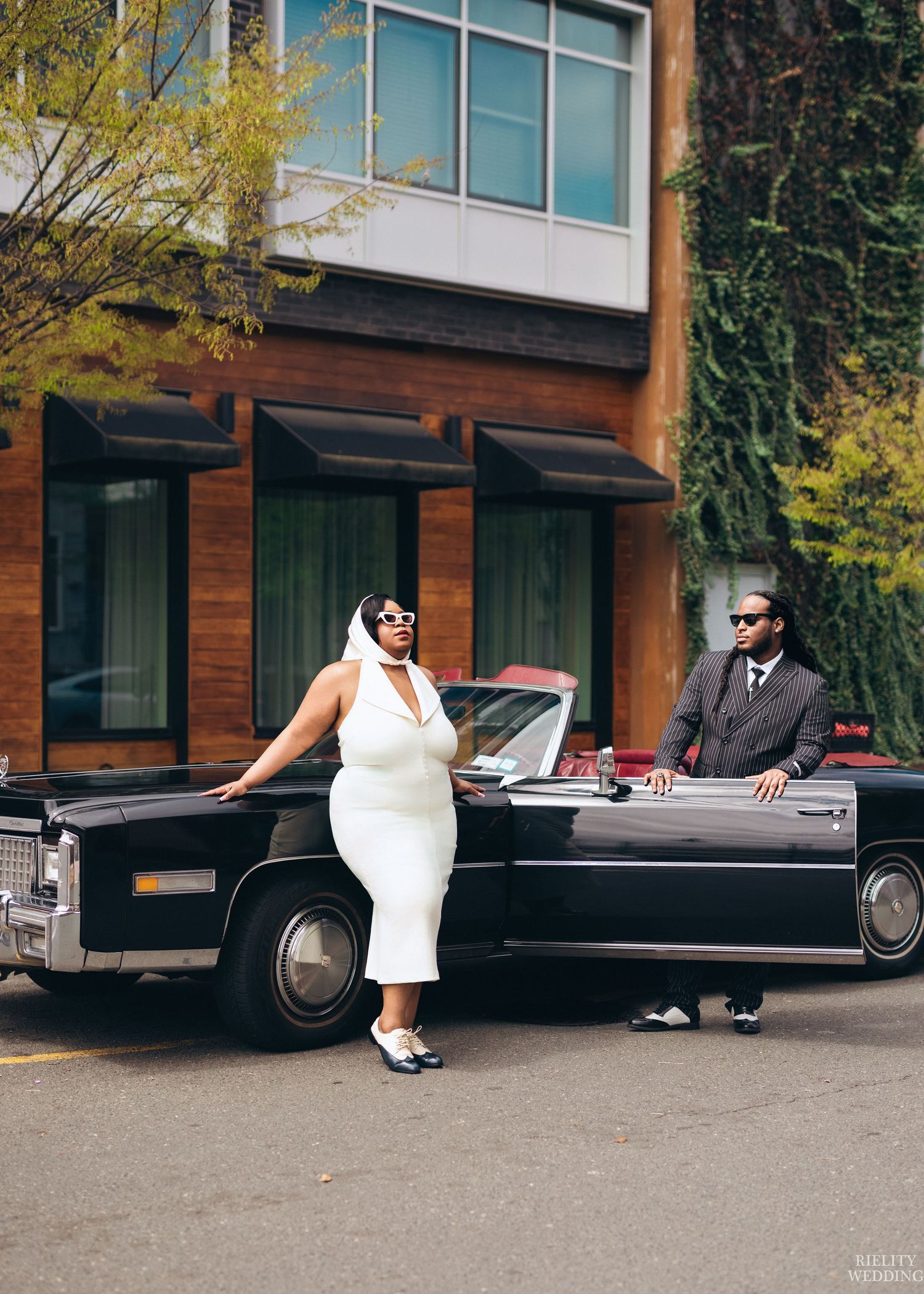 A stylish couple in formal attire poses with a classic vintage Cadillac convertible outside a modern building.