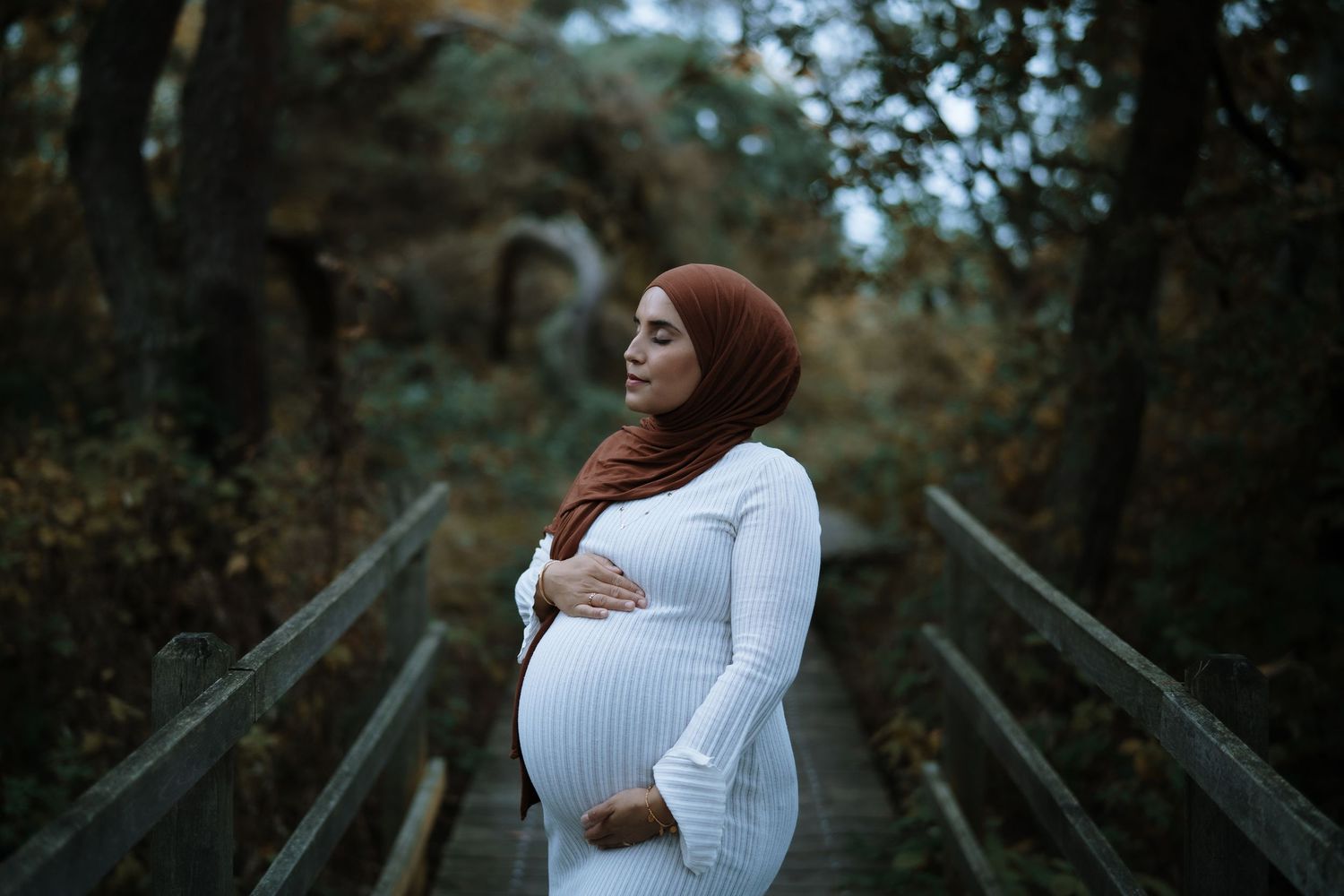 A pregnant woman in a white dress stands on a wooden bridge surrounded by lush greenery.