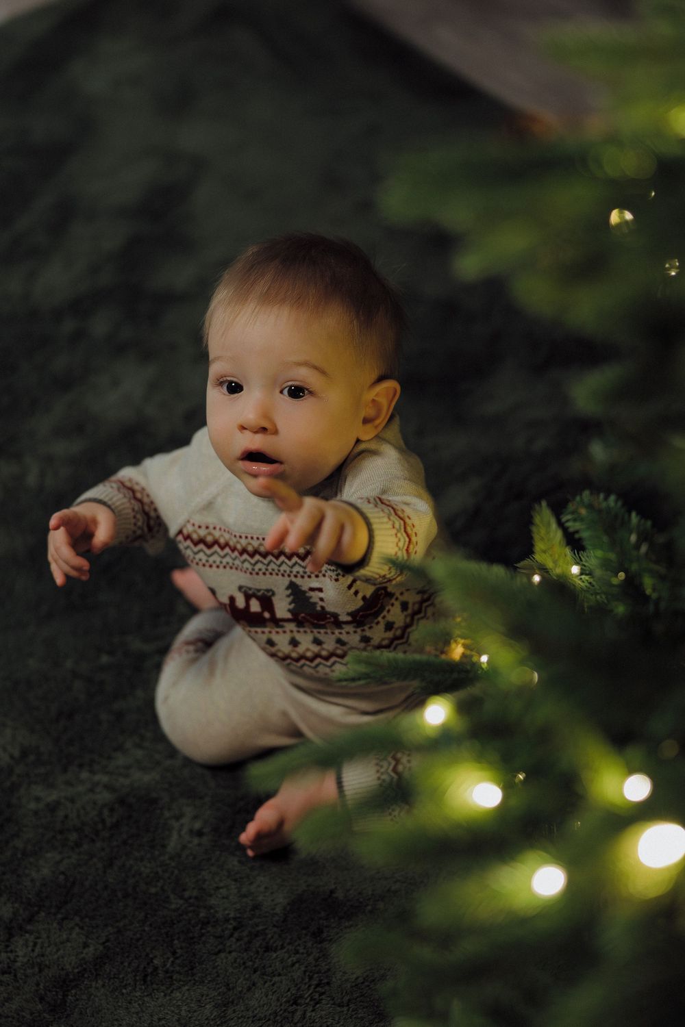 Baby in a cozy patterned sweater sits on a dark blanket near twinkling Christmas lights.
