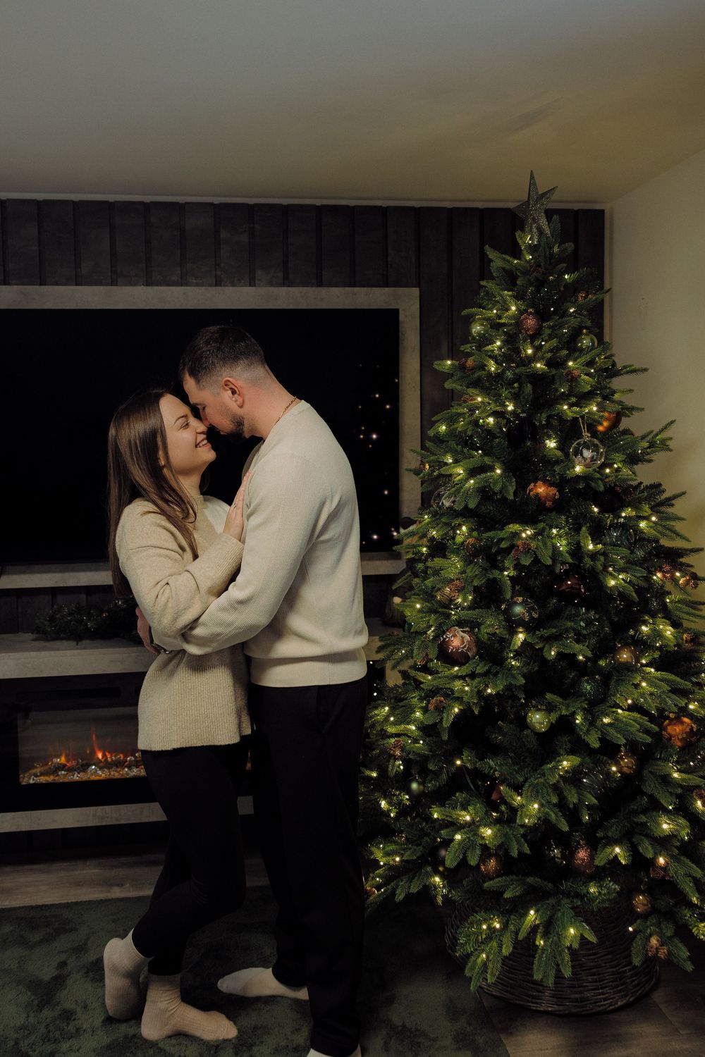 A romantic couple shares tender moments in front of a decorated Christmas tree with glowing lights and a fireplace.