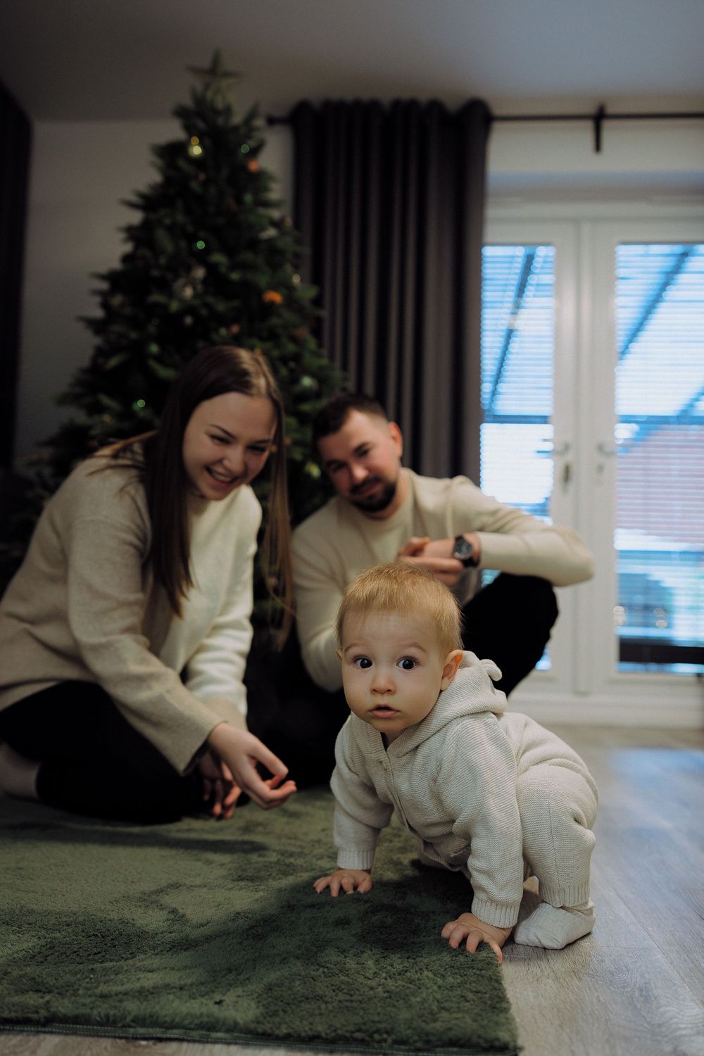 A baby in white clothing crawls on a green carpet while adults watch from behind near a Christmas tree.