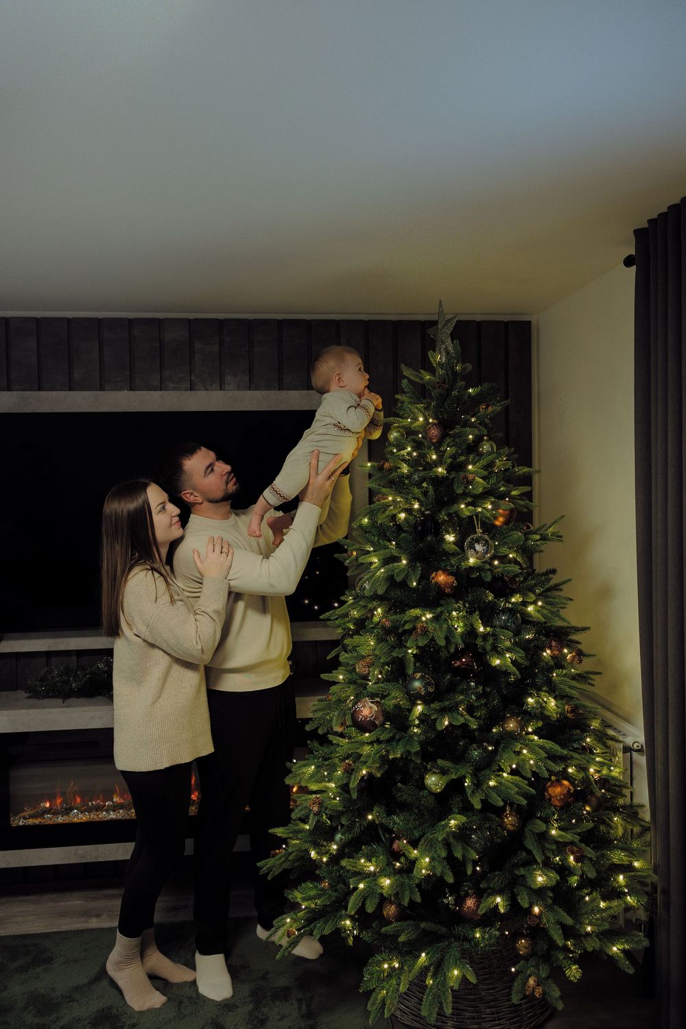 Someone decorates a Christmas tree by hanging ornaments while wearing a cream sweater and dark pants.