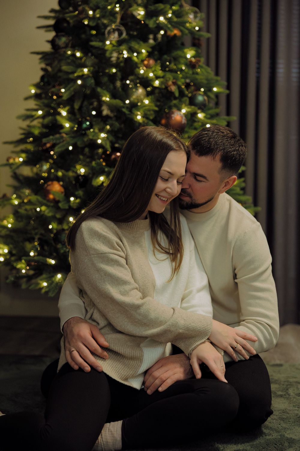 A romantic couple in matching cream sweaters shares intimate moments in front of a decorated Christmas tree.