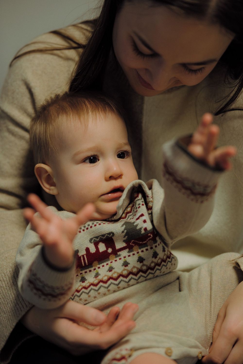 A toddler in a patterned sweater reaches out while being held by someone in a light-colored top.