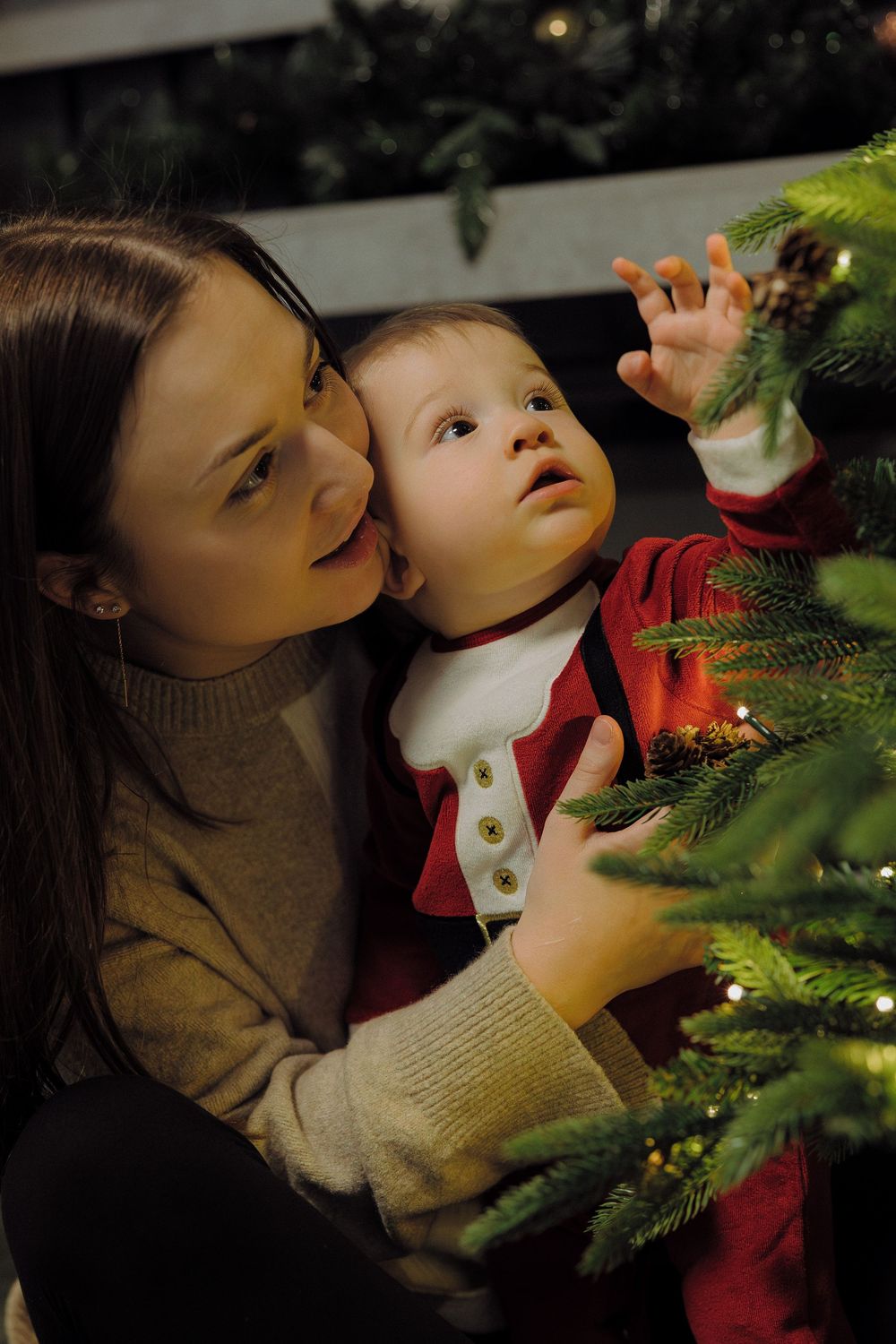 Two people share a tender moment while decorating a Christmas tree with twinkling lights and evergreen branches.