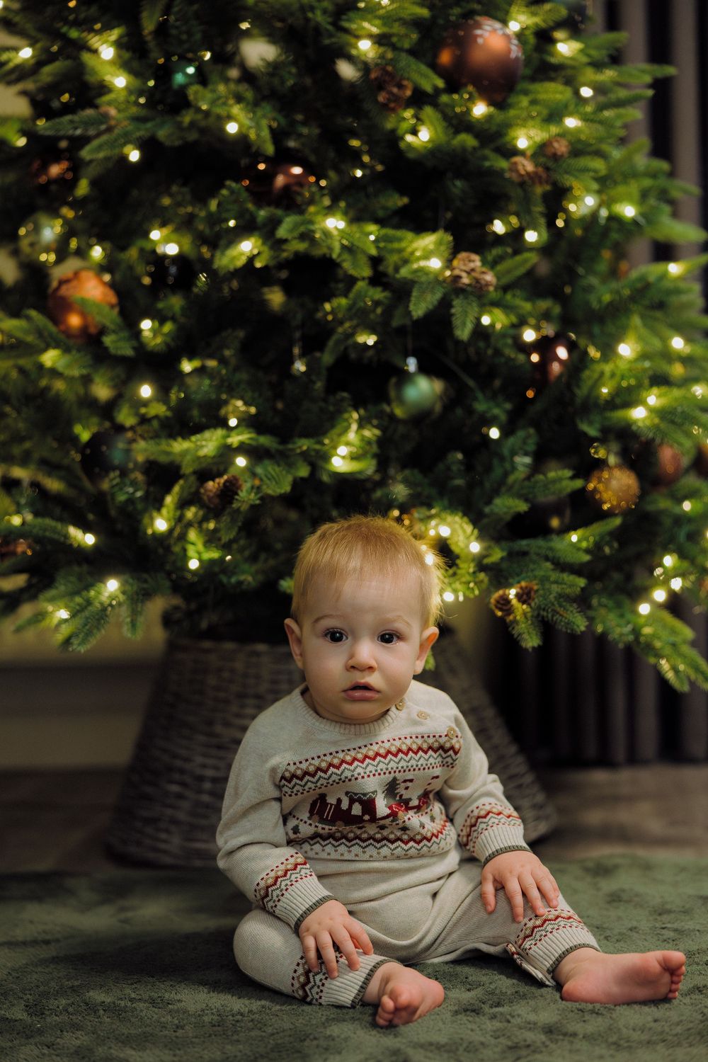 Baby in festive sweater sits beneath glowing Christmas tree with twinkling lights and greenery.