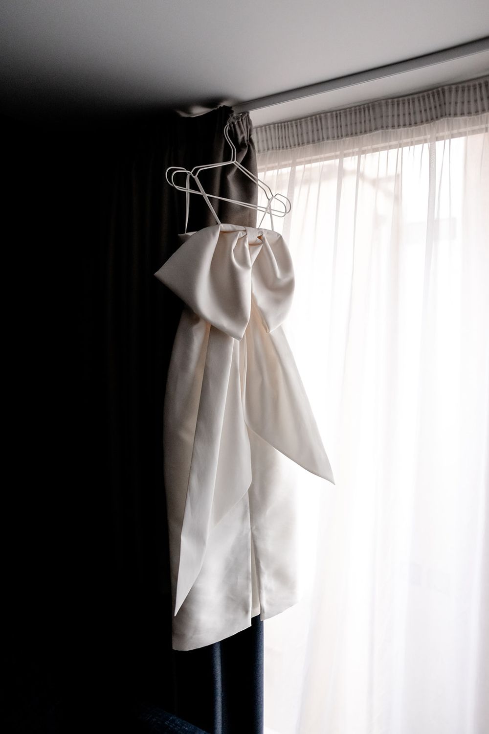 White dress shirt hanging on a hanger against a bright window with sheer curtains.