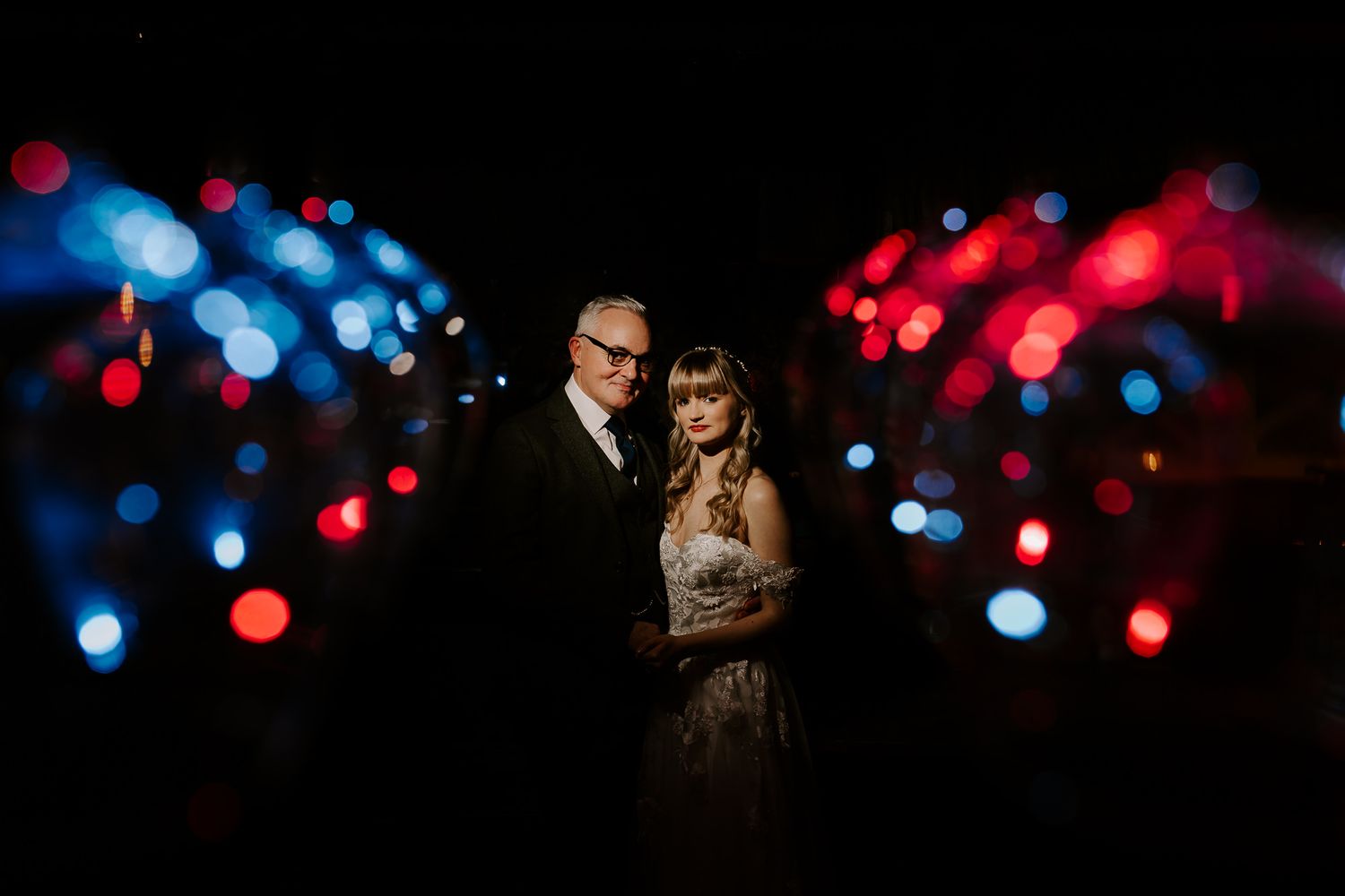 A romantic nighttime portrait of a couple embracing with red and blue bokeh lights creating a dreamy backdrop.