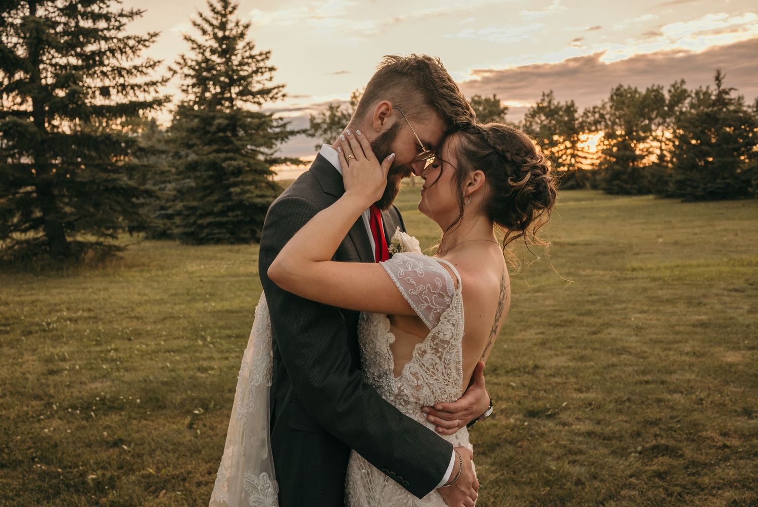 A couple shares an intimate embrace in a field at sunset with evergreen trees silhouetted in the background.