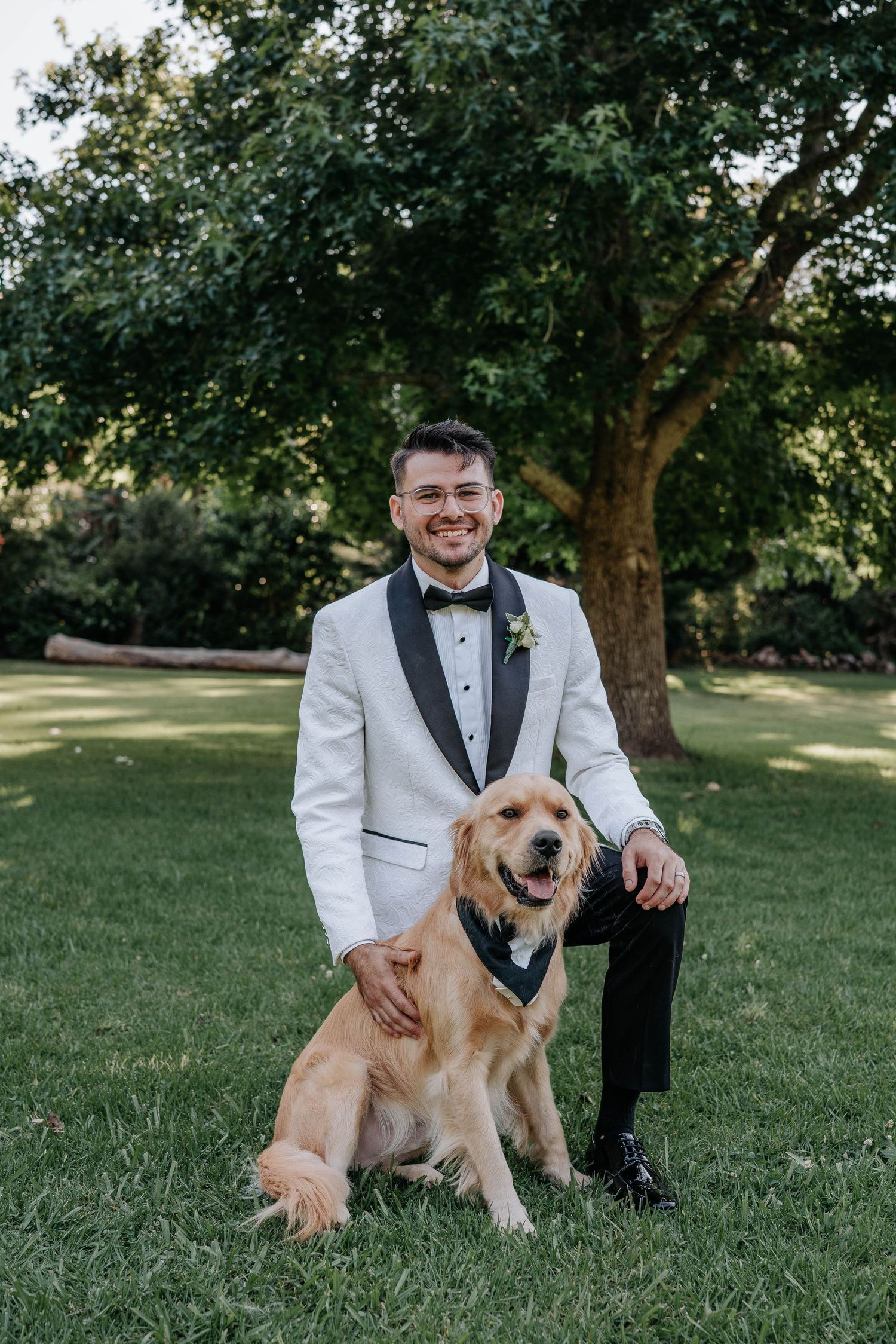 A Golden Retriever in a navy bow tie sits on grass next to someone wearing a white tuxedo jacket during a formal event.