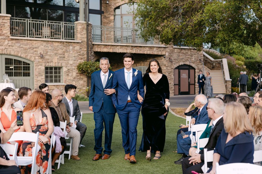 Wedding guests seated in white chairs at an outdoor ceremony in front of a rustic brick building.