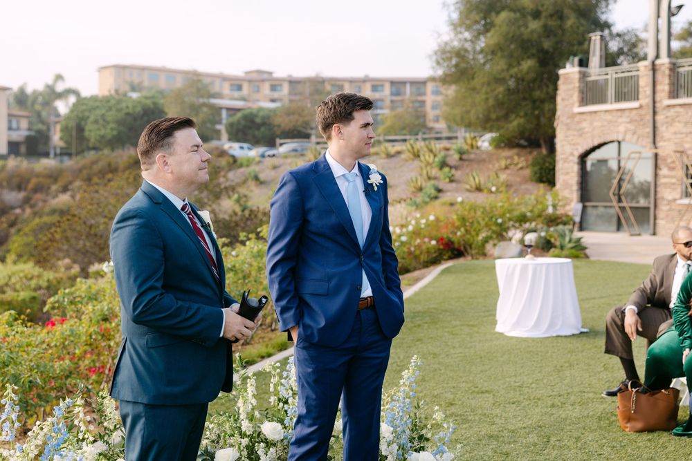 Wedding party gathers outdoors on a grassy hillside with buildings in the background during a sunny ceremony.