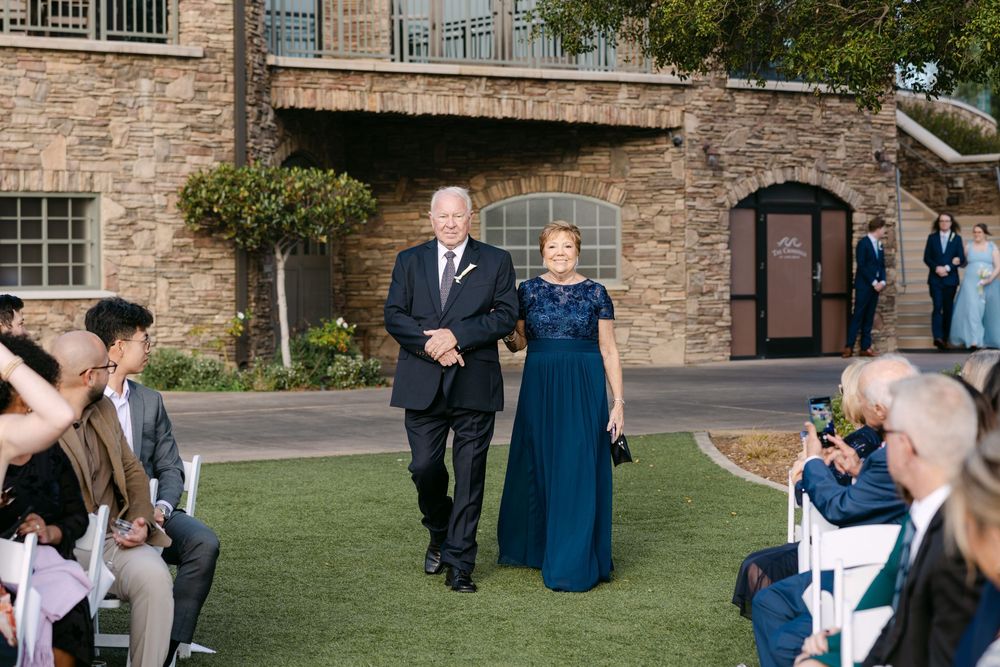 Wedding guests gather on a lawn outside a historic stone building during an outdoor celebration.