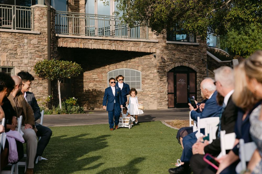 Wedding guests gather on a sunny lawn outside a brick building for an outdoor ceremony.