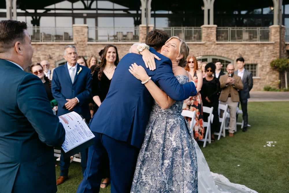 Wedding guests in navy blue attire gather outdoors on a grassy lawn near a brick building during a celebration.