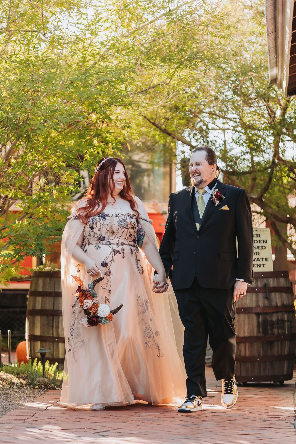A couple walks together outdoors on a brick path, with the bride wearing a blush floral gown and groom in black suit.