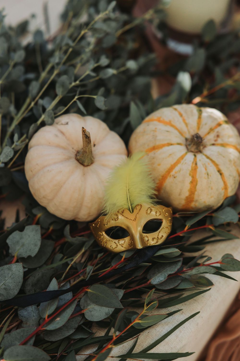 Small white decorative pumpkins arranged with dark green eucalyptus leaves and a golden masquerade mask for fall decor.