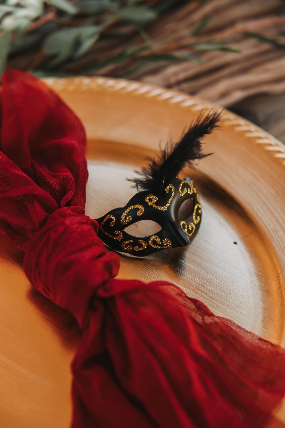 Small decorative black feathered masquerade mask with gold accents sits beside red fabric on wooden surface.