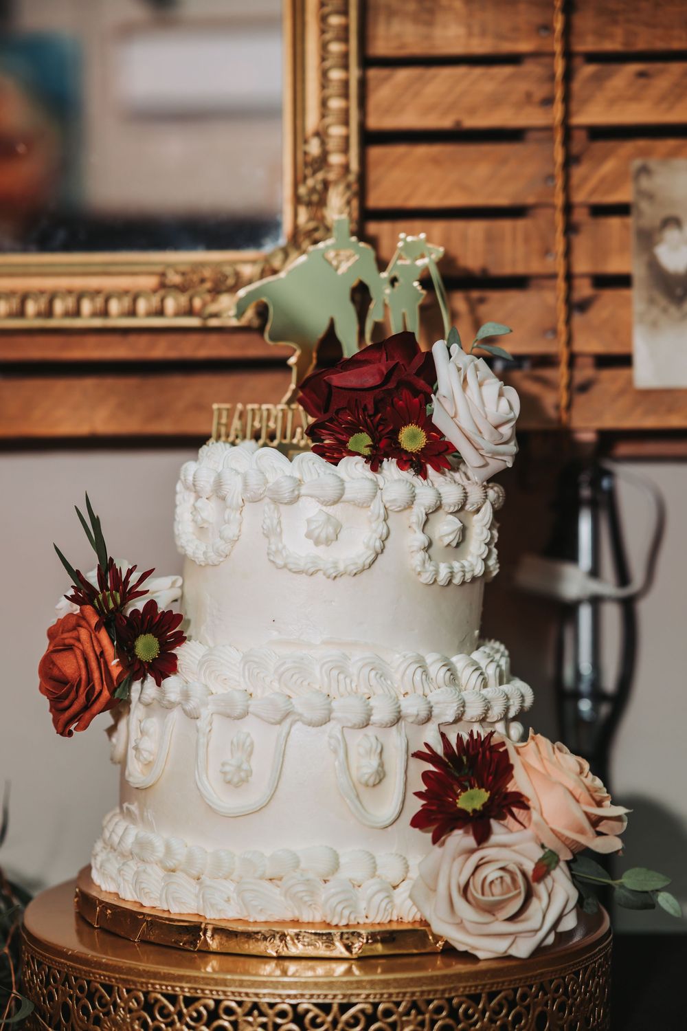 Elegant white tiered wedding cake decorated with burgundy roses and intricate piping on a gold stand.