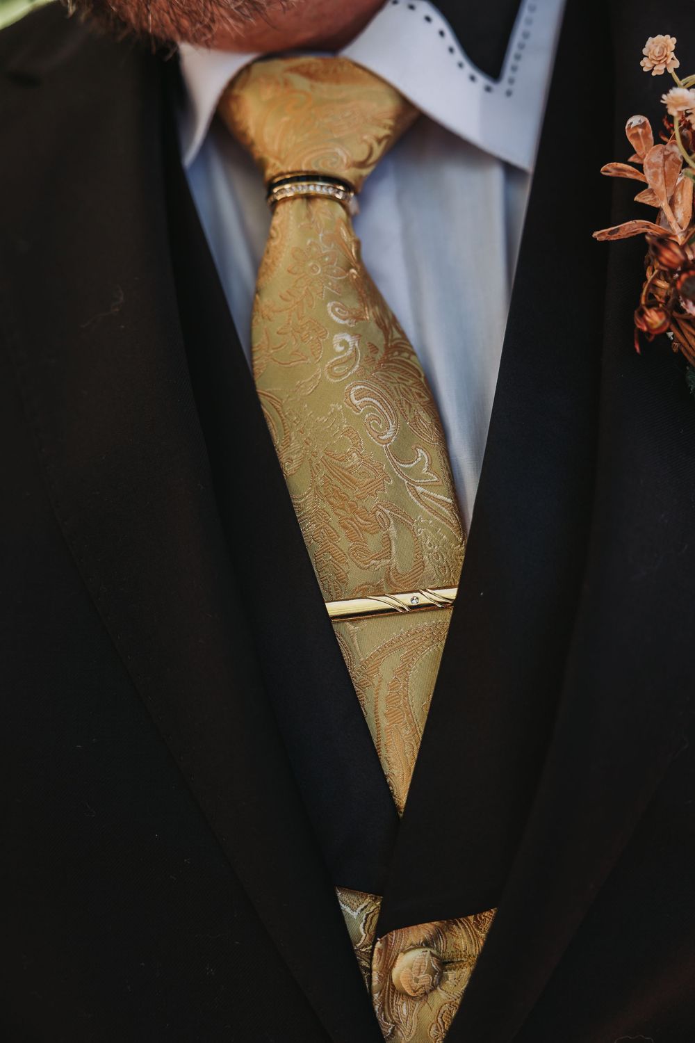 A close-up view of a gold paisley patterned tie worn with a black suit jacket and light blue dress shirt.