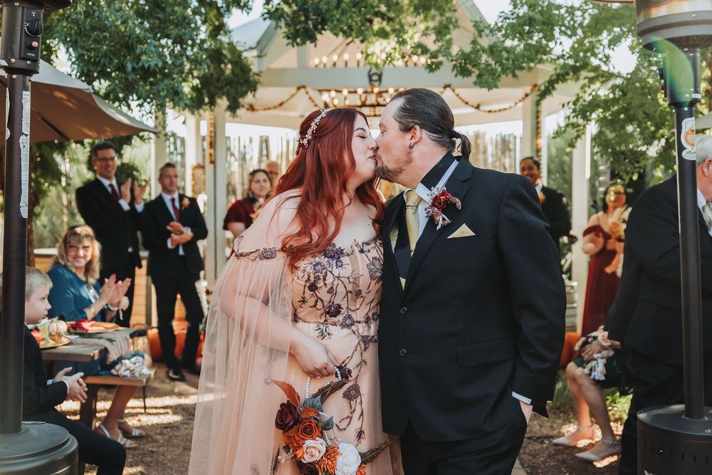 A couple shares a romantic kiss at an outdoor wedding ceremony under a gazebo with guests looking on.