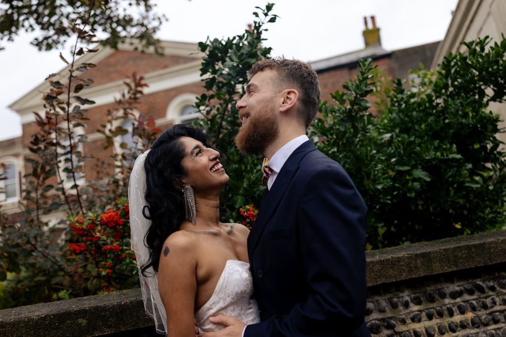 Romantic wedding couple embracing outdoors against a brick building with lush greenery.