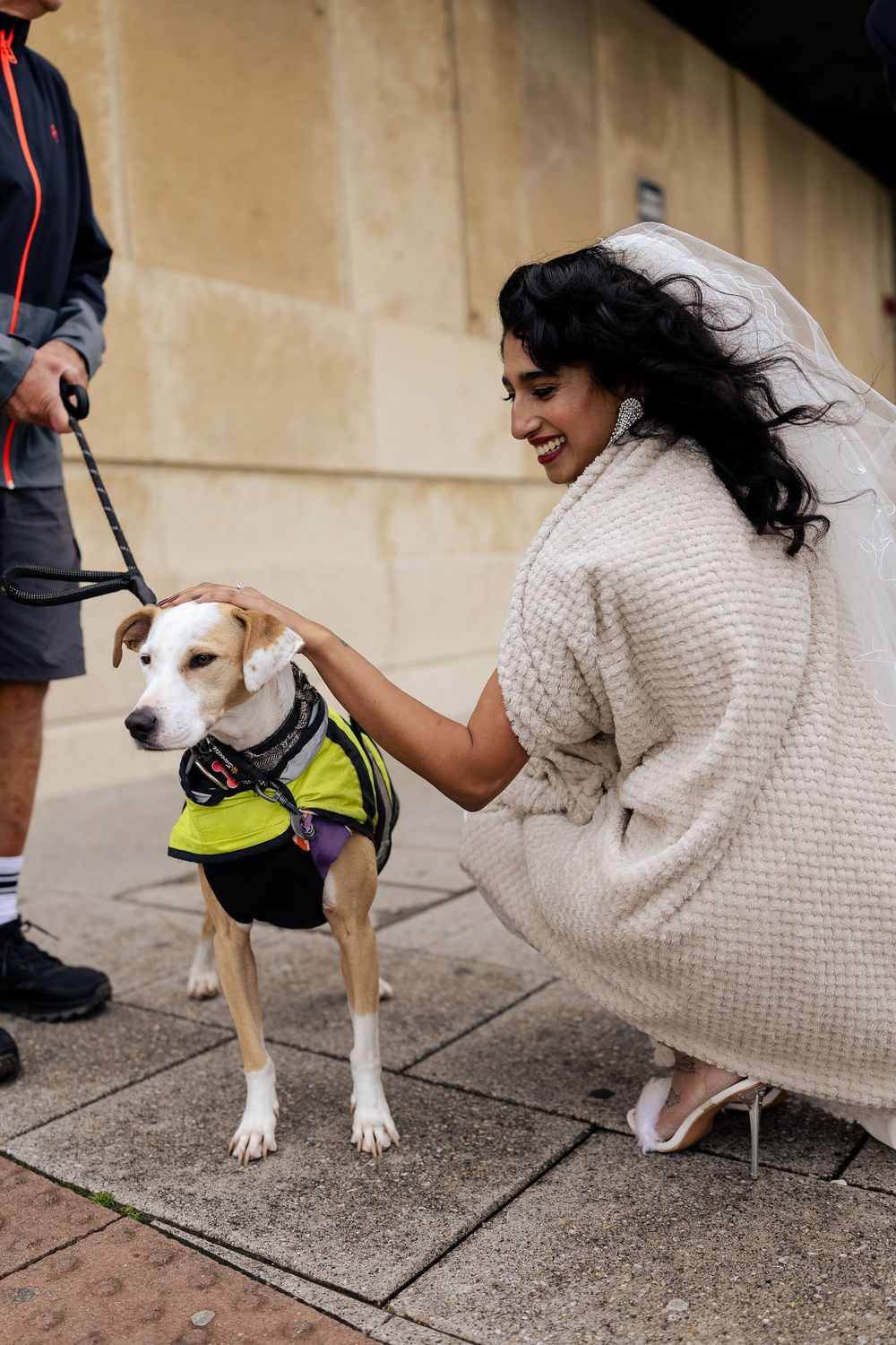 Person in white dress petting a beige dog wearing a yellow safety vest on a sidewalk.