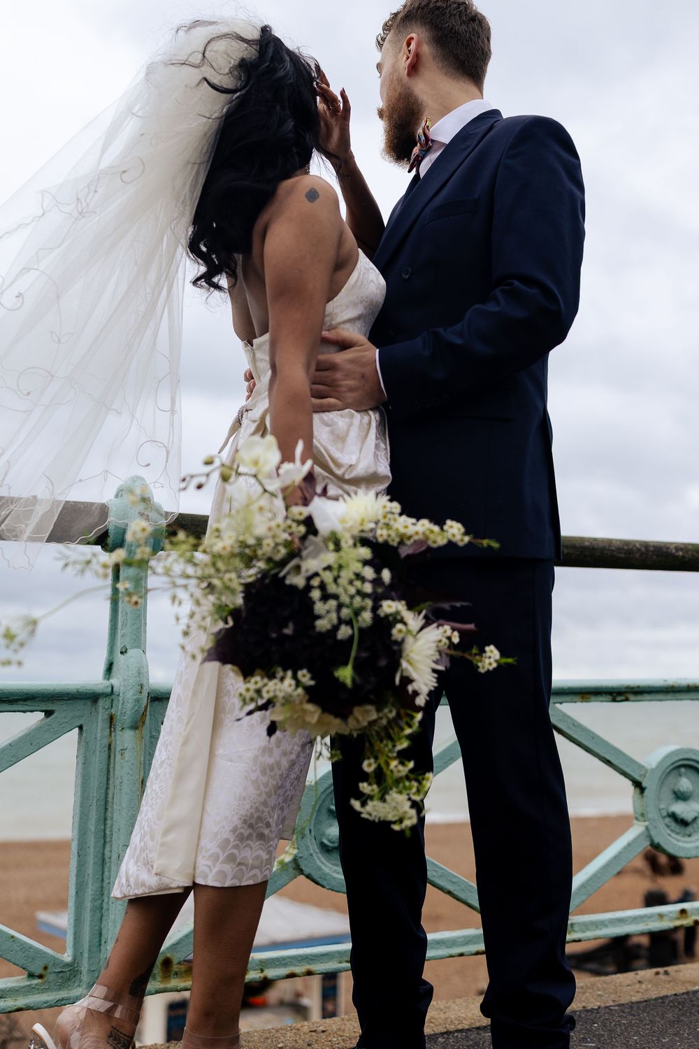 Wedding couple stands on boardwalk overlooking water with white floral bouquet.